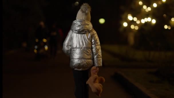 Back view of lonely little girl walking away in slow motion at night in urban city. Lost kid strolling with toy teddy bear outdoors in dusk. Social problems and childhood. — Stock Video
