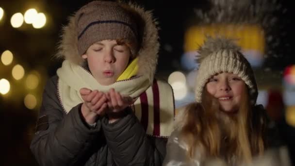 Joyeux frère et sœur caucasiens mignons soufflant et jetant des flocons de neige au ralenti au crépuscule à l'extérieur. Portrait de mignon garçon et fille joyeux profitant de la célébration du Nouvel An avec des lumières à l'arrière-plan. — Video