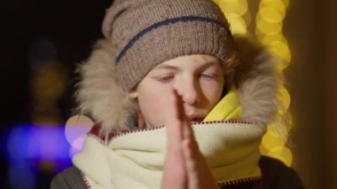 Dondurulmuş Kafkasyalı çocuğun yakın plan fotoğrafı. El ısıtan, avuçlarını ovuşturan ve açık havada esen. Yeni yıl arifesinde arka planda altın ışıklı şirin bir çocuk portresi..