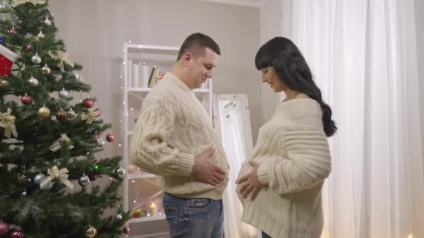 Χαρούμενος Καυκάσιος άντρας και έγκυος γυναίκα συγκρίνουν κοιλιές στο σπίτι παραμονή Πρωτοχρονιάς. Χαρούμενα αντρόγυνο που διασκεδάζουν τα Χριστούγεννα σε εσωτερικούς χώρους. Χαρά και τρόπος ζωής. — Αρχείο Βίντεο
