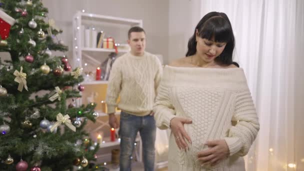 Όμορφη έγκυος καυκάσια γυναίκα χαϊδεύει την κοιλιά σε αργή κίνηση και μιλάει με θολή άνδρα που στέκεται στο παρασκήνιο σε εσωτερικούς χώρους. Χαρούμενη μέλλουσα απολαμβάνοντας την εγκυμοσύνη τα Χριστούγεννα με το σύζυγο. — Αρχείο Βίντεο