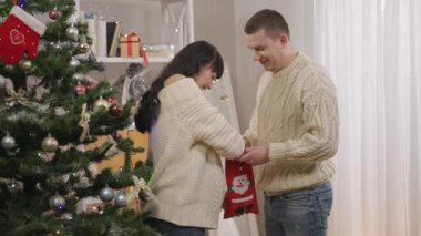 Mutlu çift evde Noel çorabından şeker alırken eğleniyor. Gülümseyen Kafkas hamile kadın ve neşeli adamın yan görünüm portresi. Yeni yıl arifesini kapalı yerde kutluyorlar..