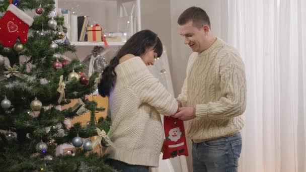 喜んで幸せなカップルは家でクリスマスの靴下からキャンディーを取ることを楽しんでいます。笑顔の白人妊婦と明るい愛の男のサイドビューポートレート新年を祝う屋内. — ストック動画