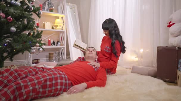 幸せな白人ブルネットの男の肖像画は、自宅でクリスマスの前夜に本を読んで美しい妊婦の膝の上に横たわっています。期待の妻と新年のお祝いを楽しむリラックスした夫. — ストック動画