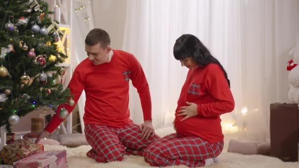 Ο χαμογελαστός σύζυγος δίνει Χριστουγεννιάτικο δώρο στην ενθουσιασμένη έγκυο γυναίκα με τις κόκκινες πιτζάμες. Ευτυχισμένος αγαπητός Καυκάσιος άνδρας και μέλλουσα γυναίκα γιορτάζει παραμονή Πρωτοχρονιάς στο σπίτι. Αργή κίνηση. — Αρχείο Βίντεο