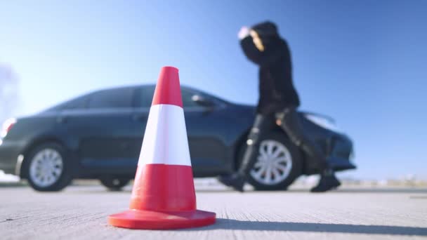 Gros plan du cône de la route avec une femme confiante floue marchant vers la voiture en arrière-plan et assise sur le siège passager. Gros plan de jeune femme caucasienne sur la route à l'extérieur. Mode de vie et voyages. — Video