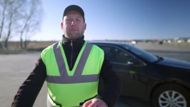 Kendinden emin beyaz araba eğitmeninin portresi el ele tutuşup kameraya bakıyor. Yeşil yelekli bir adam arabanın önünde dikiliyor. Sürüş eğitimi ve meslek konsepti.