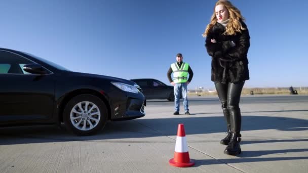 Molesto hermosa mujer delgada pateando cono de carretera con difuminado instructor automático de pie en el fondo. Aprendizaje caucásico insatisfecho que tiene problemas en los cursos de conducción. Seguridad vial y tráfico. — Vídeos de Stock