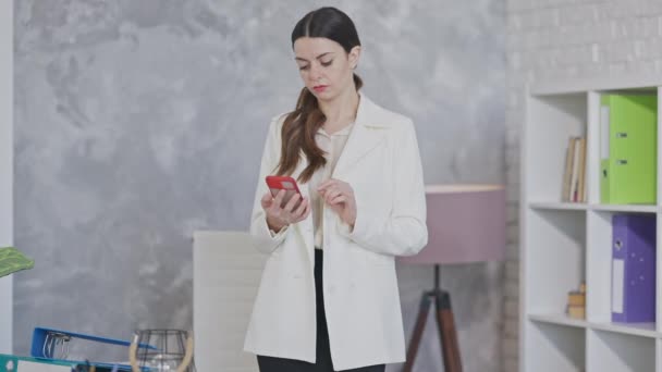 Портрет фокусованої стрункої вродливої кавказької жінки, що дивиться на смартфон. Зосереджена на успішних повідомленнях молодих бізнесменів, які стоять у кабінеті.. — стокове відео