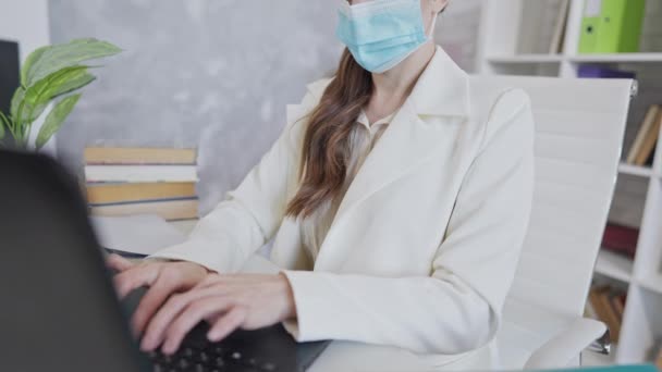 Неузнаваемая сфокусированная женщина в маске Ковида, печатающая в интернете на клавиатуре ноутбука. Уверенный тощий кавказский тысячелетие работает в офисе по борьбе с пандемией коронавируса. — стоковое видео