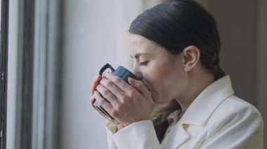 Yemek molasında çayını yudumlayan ve pencereden dışarı bakan rahat genç bir kadının yan görünüşü. Gülümseyen beyaz iş kadınının yakın plan portresi sıcak içki içip gülümsüyor. Yavaş çekim.