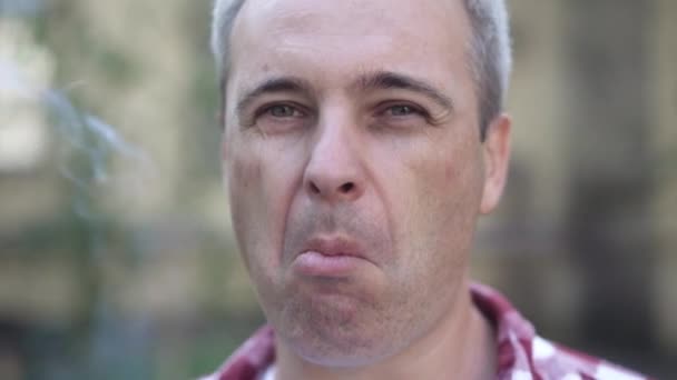 Imagen de un hombre caucásico de pelo gris fumando al aire libre soplando humo mirando a la cámara. Retrato de cerca de una persona adicta que tiene mal hábito. Adicción al tabaco. — Vídeos de Stock