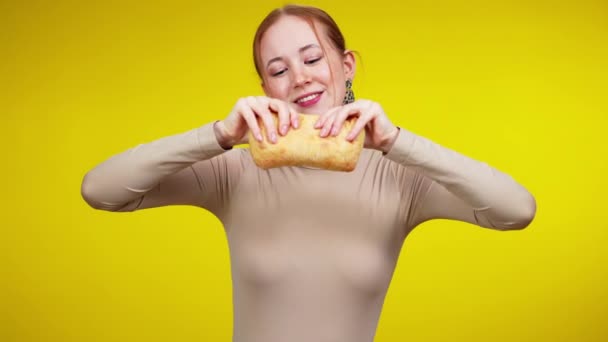 Весела усміхнена жінка розриває шибату і дивиться на камеру. Щаслива молода кавказька жінка, що тримає на жовтому тлі смачну круту пекарню.. — стокове відео
