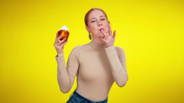 Middle shot portrait of cheerful slim young redhead woman tasting topping from small cake looking at camera smiling. Positive joyful Caucasian lady enjoying tasty sweet food at yellow background. — Stock Video