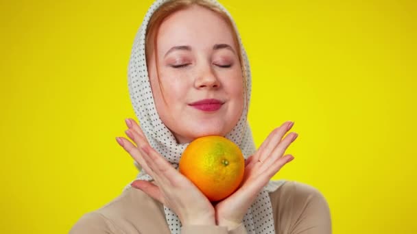 Retrato de cerca de la alegre pelirroja caucásica en pañuelo sosteniendo fruta naranja mirando a la cámara sonriendo. Joven linda dama posando en el fondo amarillo con dulce sabrosa comida. — Vídeo de stock