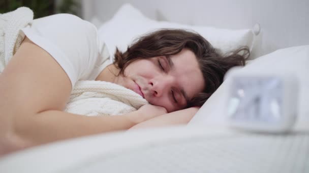 Close-up van slapende blanke man die wakker wordt, wekker uitzet en zich verstopt onder een witte deken. Portret van een slaperige jonge brunette in bed in de ochtend. Vermoeidheid en rustbegrip. — Stockvideo