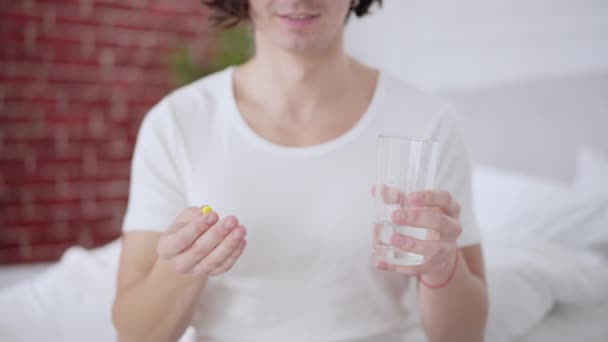 Αρσενικά χέρια κρατώντας κίτρινο χάπι και ένα ποτήρι νερό με αγνώριστη θολή χαμογελαστός άνθρωπος στο παρασκήνιο. Νεαρός Καυκάσιος που ποζάρει στο υπνοδωμάτιο με αντικαταθλιπτικά ή βιταμίνη το πρωί. — Αρχείο Βίντεο