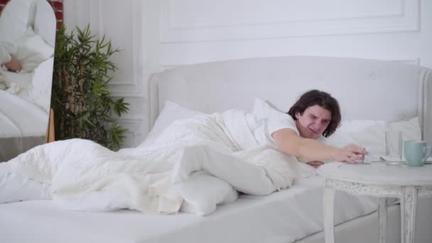 Szerokie ujęcie smutnego niezadowolonego mężczyzny wyłączającego budzik i zamykającego głowę poduszką. Portret nieszczęśliwego białego tysiąclecia rano w domu. Przebudzenie i styl życia. — Wideo stockowe
