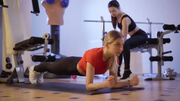 Mulher jovem esbelta concentrada em pé na posição de prancha no tapete de exercício com millennial magro borrado usando halteres para treinamento de bíceps no fundo. Treinamento de esportistas caucasianos confiantes. — Vídeo de Stock