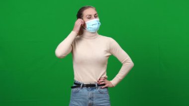 Esmer, beyaz bir kadın Covid-19 maskesini çıkarıyor. Kameraya bakıp iç çekiyor ve gülümsüyor. Koronavirüs virüsü salgınında yeşil ekranda poz veren kendine güveni zayıf kadının portresi.
