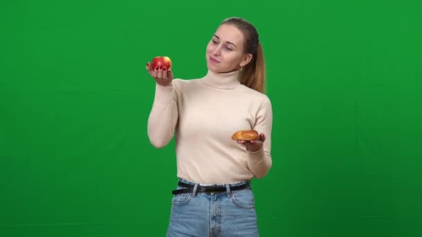Тонкая женщина выбирает здоровое яблоко, держа в руках фрукты и булочку. Портрет молодой белой женщины, выбирающей еду на зеленом экране. Концепция здорового и нездорового питания. — стоковое видео