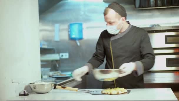 Retrato de cocinero masculino profesional concentrado en máscara facial Covid-19 y guantes que agregan ingredientes en la pizza en la cocina. Hombre caucásico trabajando en pizzería en pandemia de coronavirus. — Vídeos de Stock
