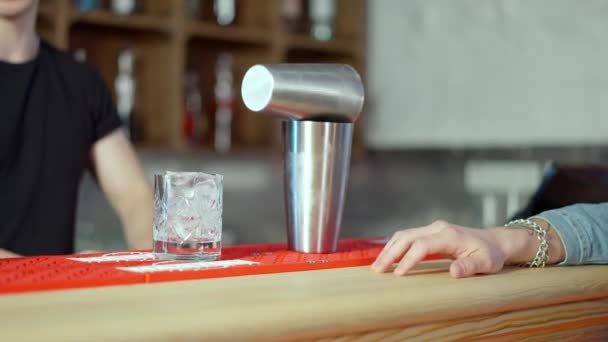 Agitatore da cocktail e bicchiere con ghiaccio in piedi sul bancone del bar con barista sfocato che lavora sullo sfondo e mano maschile che bussa le dita. Barista professionista che prepara bevande per il cliente nel pub. — Video Stock