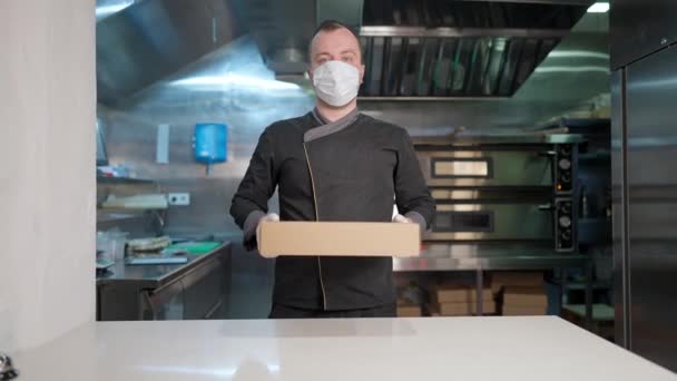 Портрет уверенного кавказского повара в маске Ковида, растягивающего бумажную коробку с заказом еды на вынос на камеру. Профессиональный кавказский повар, позирующий на кухне ресторана в связи с пандемией коронавируса. — стоковое видео