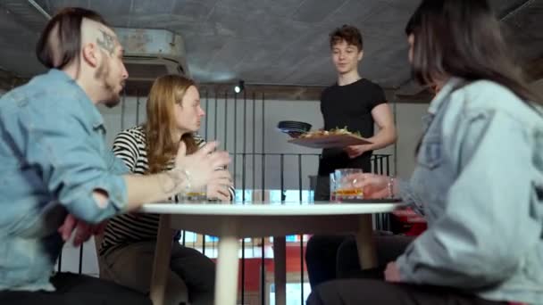 Kelner przynoszący pizzę dla grupy wesołych białych przyjaciół siedzących przy stole w kawiarni. Szerokie ujęcie pozytywnego młodzieńca serwującego spokojne, beztroskie spotkanie milenium w pizzerii. Serwis i styl życia. — Wideo stockowe