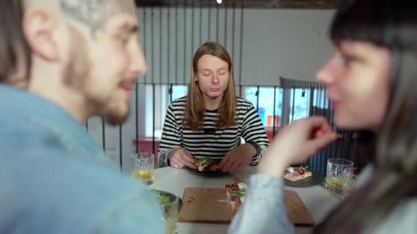 Размытая молодая пара разговаривает, как смешной человек ест пиццу на заднем плане. Позитивный беззаботный кавказский мужчина с длинными волосами с удовольствием дегустирует вкусную еду в пиццерии, сидя с друзьями. — стоковое видео