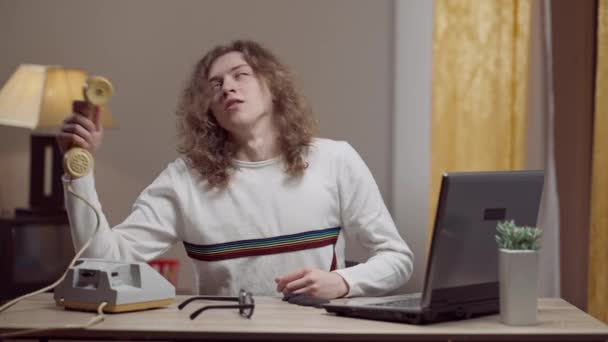 一个年轻的复古男人拿起电话笑着说话的画像。80年代90年代，白种人长发的中景男子坐在餐桌旁，带着老式电话和笔记本电脑. — 图库视频影像