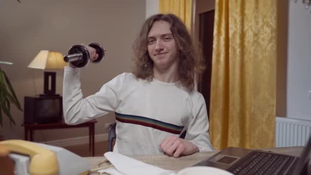 Zelfverzekerde tachtiger jaren man zit aan tafel met retro telefoon en laptop te zien op halter in de hand en glimlachend kijkend naar de camera. Portret van zelfbewuste blanke man binnen in huis in de jaren negentig. — Stockvideo