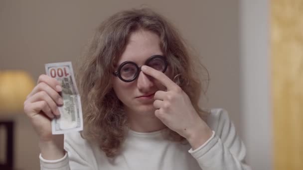 Retrato de un hombre confiado e inteligente ajustando anteojos mostrando que tengo una idea gesto sosteniendo billete de dólar. Éxito joven caucásico nerd de 1980 posando en el interior de casa. Inspiración y éxito. — Vídeos de Stock