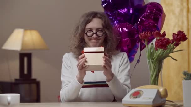 Střední snímek pozitivního retro nerda natahujícího dárkovou krabici na kameru a nastavujícího brýle. Portrét usmívajícího se mladého bělocha brunetky, který dává dárek na dovolené v 80. nebo 90. letech. — Stock video