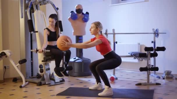 젊고 자신감 있는 여성이 쪼그려 앉아서 운동 기구를 사용하여 배경 작업을 하는 것을 바라보는 측면이다. 두 명의 날씬하고 매력적 인 여자 가 체육관에서 운동하는 모습. — 비디오