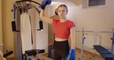 Kendine güvenen kadın boksörün portresi kameraya bakıyor ve güçlü bir hareket sergiliyor ve boks eldiveniyle pazılara vuruyor. Spor salonunda poz veren zayıf, güçlü beyaz kadın. Sinema 4k ProRes Merkezi.