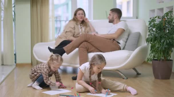 Wide shot of relaxed happy little girls drawing with blurred couple of parents sitting on couch talking at background. Carefree Caucasian family enjoying weekend at home indoors. Lifestyle concept. — Stock Video