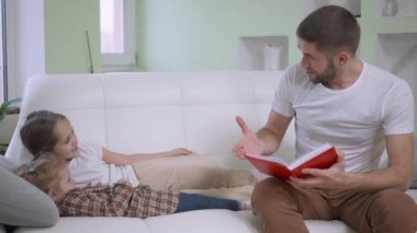Sakallı esmer baba, neşeli beyaz kızlara kitap okuyor ve kanepede gülüyor. Beyaz bir adamın güzel kızlarıyla hafta sonunu evde geçirmesinin portresi. Boş zaman ve birlik.