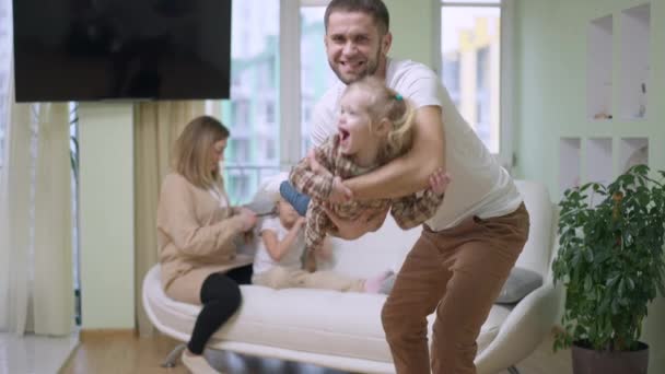 Radosny ojciec kołyszący małą dziewczynką trzymającą dziecko w rękach. Pozytywnie śmiejący się biały mężczyzna bawiący się z córką w zwolnionym tempie. Zamazane kobieta i dziecko siedzi w tle na kanapie w salonie. — Wideo stockowe