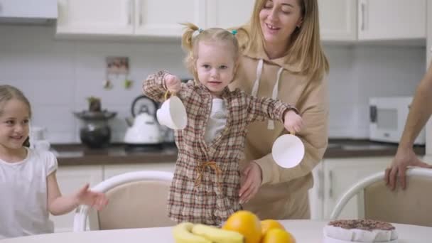 Affascinante bambina bionda che tiene le tazze in piedi sulla sedia in cucina con madre e sorella allegre che proteggono il bambino. Ritratto di bambino caucasico felice a casa con la famiglia. — Video Stock