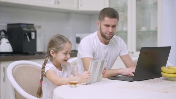 Χαρούμενη ευτυχισμένη κοπέλα γελώντας βλέποντας κινούμενα σχέδια στο tablet ως απασχολημένος άνθρωπος surfing Internet στο laptop στο παρασκήνιο. Θετική Καυκάσια κόρη κάθεται στην κουζίνα με τον πατέρα στο σπίτι. Σύγχρονος τρόπος ζωής. — Αρχείο Βίντεο