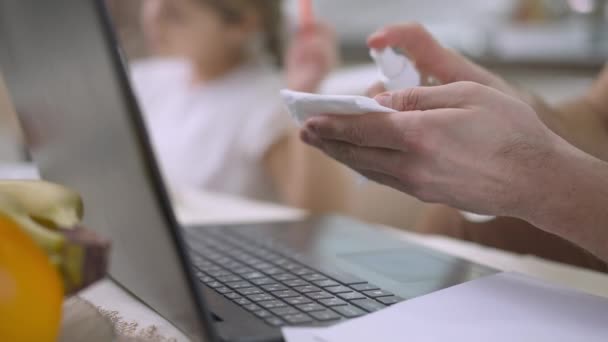 Close-up of male hands disinfecting laptop keyboard with sanitizer as blurred little girl drawing at background. Ayah Kaukasia yang tidak dikenal bekerja secara online dari rumah dengan putri yang beristirahat di rumah. — Stok Video