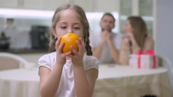 Wesoła dziewczyna żonglująca pomarańczą i pachnąca pysznymi owocami patrząca na uśmiechniętą kamerę. portret szczęśliwego, białego dziecka pozującego ze zdrowym deserem i rozmazaną parą rodziców w tle — Wideo stockowe
