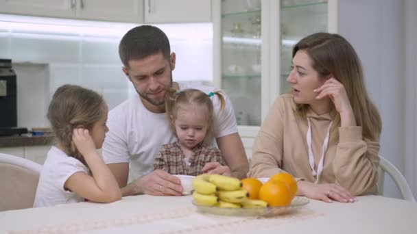 Χαλαρωμένη οικογένεια Καυκάσιων κάθεται στο τραπέζι στην κουζίνα μιλώντας κοιτάζοντας την κάμερα και χαμογελώντας. Πορτρέτο του ευτυχισμένου άνδρα γυναίκα και τα κορίτσια θέτουν στο σπίτι το πρωί. Έννοια ενότητας και αναψυχής. — Αρχείο Βίντεο