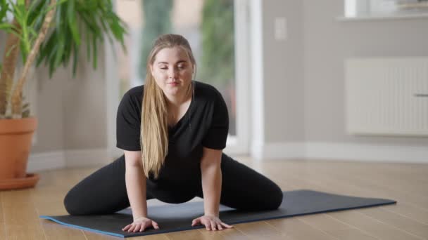 Visão frontal retrato de encantador sorridente desportista plus-size olhando para câmera respirando em pose de cordel no tapete de exercício. Motivado confiante caucasiano obeso mulher formação dentro de casa. — Vídeo de Stock