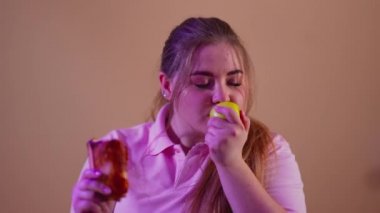 Aşırı kilolu, elma kokan ve sağlıksız yüksek kalorili sandviç yiyen komik genç bir kadının yakın plan portresi. Beyaz tenli, tombul bir kadın içeride abur cubur yiyor. Seçim ve obezite.