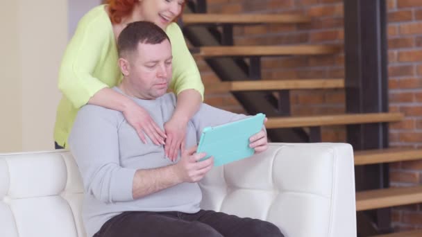 Istri yang ceria dan penuh kasih memeluk suami yang terserap melalui internet di tablet. Wanita cantik Kaukasia Positif merangkul pria tampan santai duduk di sofa di rumah. Waktu luang keluarga. — Stok Video