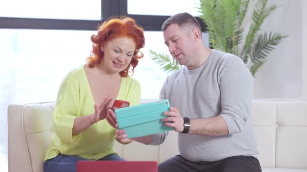 Pareja caucásica adulta positiva navegando por Internet hablando y sonriendo sentada en el sofá en casa. Retrato de hombre y mujer feliz y amoroso disfrutando de ocio de fin de semana en interiores. Concepto de estilo de vida. — Vídeo de stock