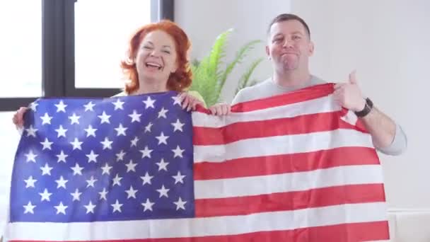 Orgulhoso casal adulto posando com a bandeira americana mostrando o polegar para cima e olhando para a câmera com o sorriso dos dentes. Retrato de homem e mulher caucasianos parciais com símbolo nacional em casa dentro de casa. — Vídeo de Stock