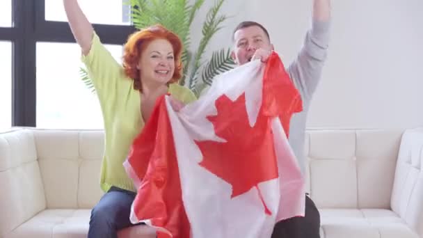 Gelukkig stel immigranten met Canadese vlag die volkslied zingen op de bank in de woonkamer. Video chat POV van vrolijke gelukkige man en vrouw die genieten van immigratie naar Canada. — Stockvideo
